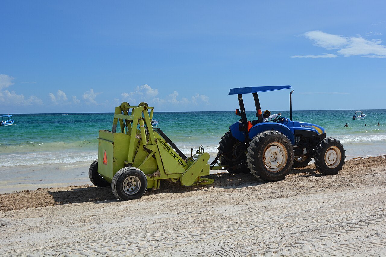 A tractor towing a beach-cleaning machine across the sand at Tulum with turquoise water behind
